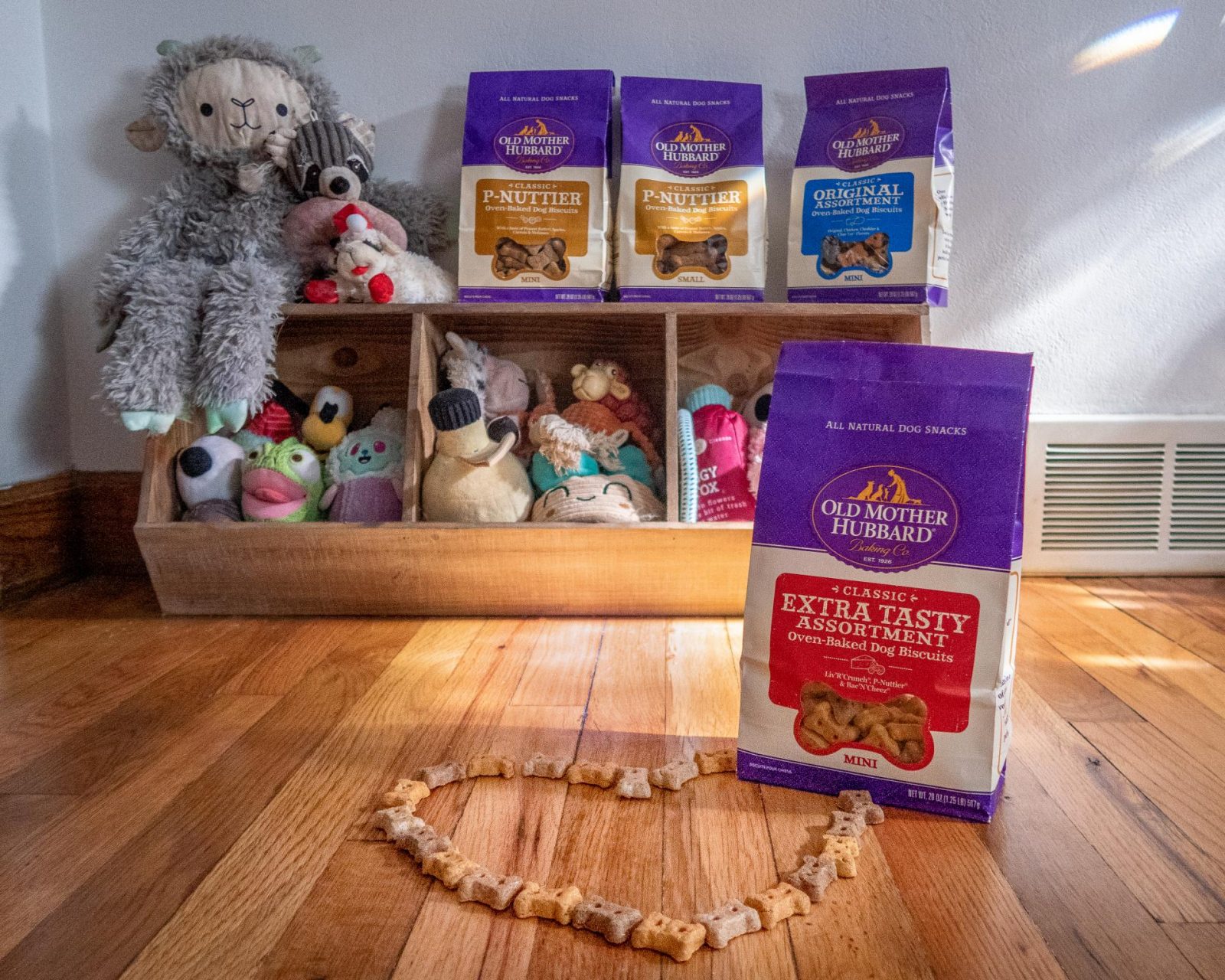 Old Mother Hubbard dog treats are lined up on Maisie the Saint Bernard's toy box, with the Extra Tasty Assortment bag in focus in front of the photos.